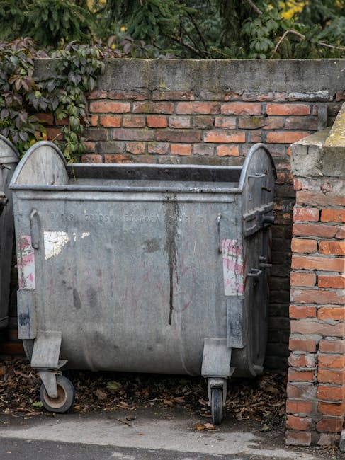 A large, grey metalwheelie bin situated outside a brick wall and a concrete wall, positioned on a paved area next to some soil and sparse vegetation. The bin has visible signs of wear, including rust and faded stickers, and features small wheels at the bottom for mobility. It is placed adjacent to a brick pillar and a doorstep, indicating its location at the entrance of a residential property. The environment appears to be outdoors in an urban or suburban setting, with natural light illuminating the scene. This image relates to house removals and waste disposal services, exemplifying the collection of bulky waste before or during a home relocation process, as offered by Man with Van Hither Green.