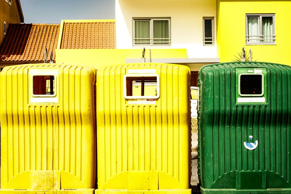 Two yellow wheelie waste bins and one green wheelie waste bin positioned outside a residential property in front of a modern building with white and yellow walls and a red tiled roof. The bins are made of ribbed plastic with small square windows on the lids; the yellow bins are adjacent to each other on the left, with the green bin to their right. All bins are placed on a paved area, and the background features a contemporary building with two small balconies, each with a sliding glass door and a metal railing, under bright daylight. This scene depicts typical waste disposal containers used for bulky waste collection, relevant to house removals and waste disposal services like those offered by Man with Van Hither Green, supporting the topic of disposing bulky waste in SE13 without extra fees.