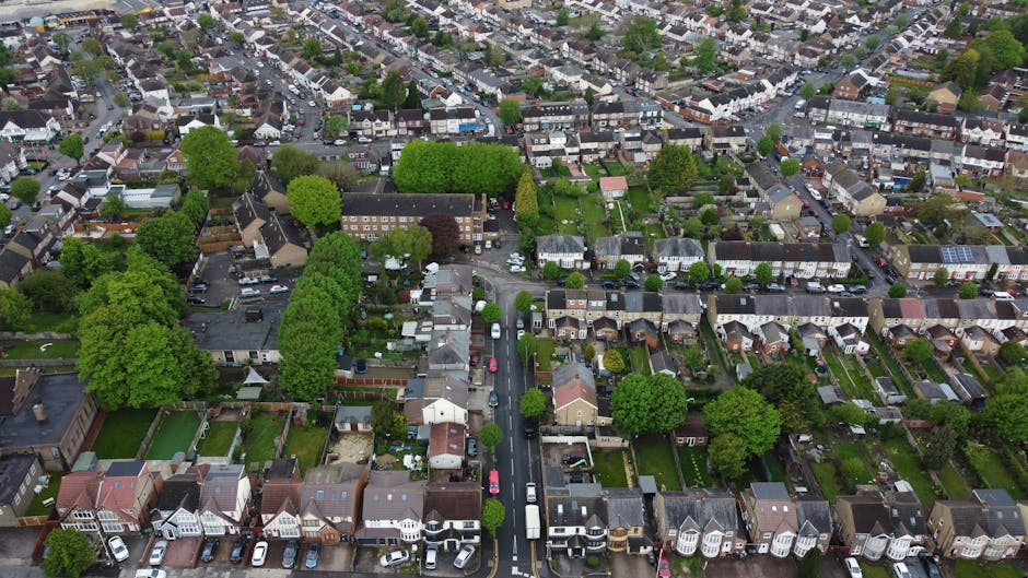 Aerial view of a densely populated residential neighbourhood in Hither Green, showing rows of terraced and semi-detached houses with pitched roofs, small gardens, and driveways. Some houses have vehicles parked in front, while others are situated on narrow streets lined with trees and greenery. Several houses are equipped with solar panels on their roofs. The area includes communal green spaces with mature trees, and the scene captures a typical home relocation environment with houses tightly packed together. The image reflects the overall setting for furniture transport and moving logistics, illustrating the environment where house removals and packing services by Man with Van Hither Green often take place.