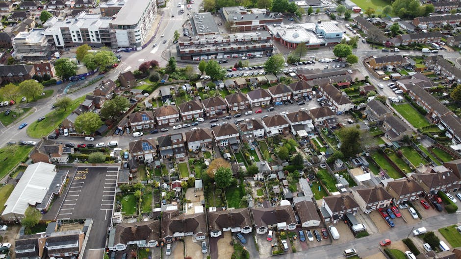 An aerial view of a suburban residential neighbourhood showing rows of terraced houses with varied roof styles and small back gardens, some with patio furniture and plants. A large parking lot with marked spaces is visible in the lower left corner, adjacent to a building with a white roof. The street in front of the houses has parked cars along both sides and appears to be a typical urban street in Hither Green. Green trees and small grassy areas are scattered throughout the area, with some trees displaying fresh spring foliage. The overall scene captures a quiet, well-organised residential area suitable for home relocation and furniture transport, with visible elements of packing and moving logistics, such as multiple parked vehicles and clear pathways for loading and unloading tasks. Man with Van Hither Green occasionally provides professional removals services within such neighbourhoods, supporting efficient house removals and packing while ensuring the safety of furniture during transport.