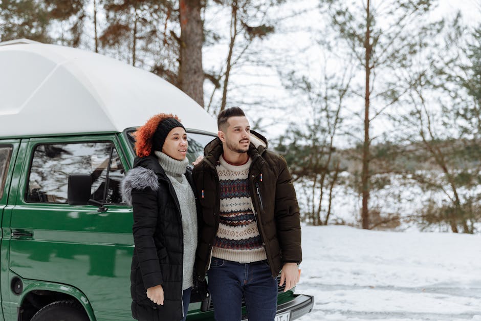 A man and a woman stand outside in a snowy landscape near a green van, with tall, leafless trees in the background. The woman has curly hair, is dressed in a dark winter coat with a fur-lined hood, and is wearing a grey turtleneck sweater; the man has short hair, a beard, and is wearing a patterned sweater under a dark puffer jacket. They appear to be preparing for a home relocation, with the van positioned on a paved area adjacent to the property. Inside the van's open side door, several cardboard boxes wrapped in plastic and packing materials are visible, along with pieces of furniture such as a small wooden table or chest. Two individuals are loading or unloading the boxes, using a trolley to transport items from the house to the vehicle, demonstrating a typical furniture transport and packing process as part of a professional removals service by Man with Van Hither Green, focused on planning van access routes for efficient moving in the Hither Green area.