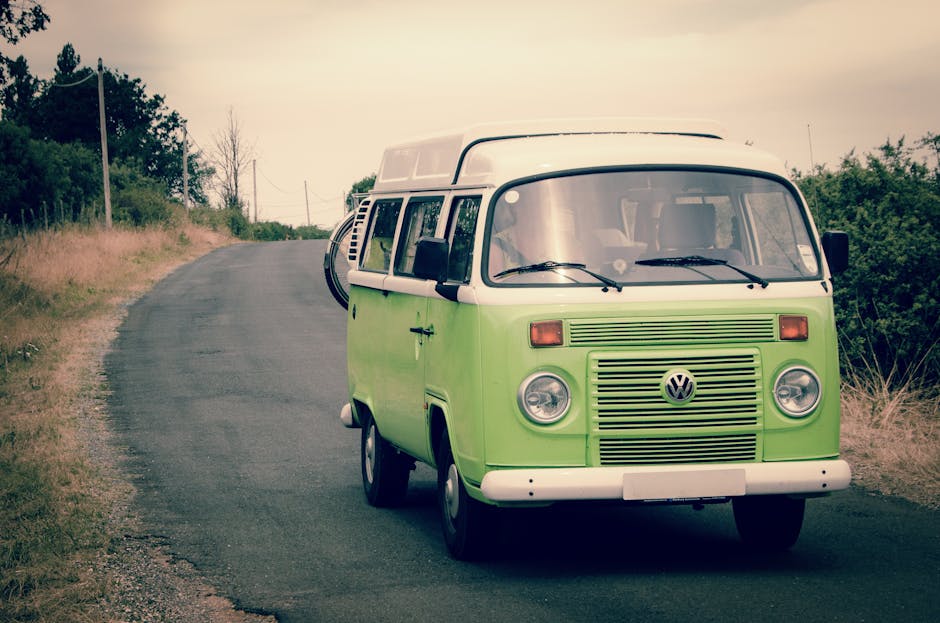 A vintage green and white Volkswagen camper van parked on a narrow rural road with a slight incline, surrounded by grassy verges and sparse trees, under an overcast sky. The vehicle is positioned close to the right side of the road, with the driver's side facing the camera. The van's front features round headlights, a prominent VW emblem, and a horizontal grille. The side windows are visible, with the sliding door closed. In the background, the road continues uphill with utility poles and power lines running parallel, and dense greenery on either side. This scene depicts a classic vehicle likely used for home relocations or furniture transport during packing and moving activities, consistent with services provided by Man with Van Hither Green, especially when considering route planning and van access for house removals on Hither Green Lane.