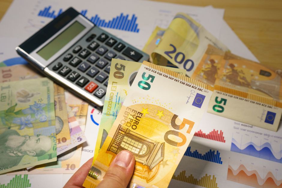 Close-up image showing a yellowish banknote placed on and partially covering three small black foam house-shaped models, which are arranged in a row. The foam houses are positioned on a surface near a