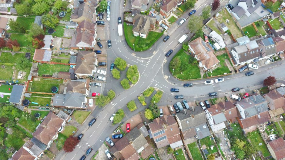 An aerial view of a residential neighbourhood showing narrow streets lined with detached and semi-detached houses, each with small front and rear gardens. Several cars are parked along the curbs, and a few vehicles are in motion on the roads. Tree-lined verges are visible, with some trees having red and green foliage. The streets intersect at a roundabout with small landscaped green spaces and trees in the center. The houses have tiled roofs, some with solar panels, and multiple properties display visible driveways or front yards. On the pavement next to a house, there are cardboard boxes, plastic wrapping, and a few wrapped furniture items, indicating a home relocation in progress. The scene is well-lit with natural daylight, capturing the routines involved in household packing and furniture transport, which are part of the moving process managed by [COMPANY_NAME], specializing in removals and house moving services.