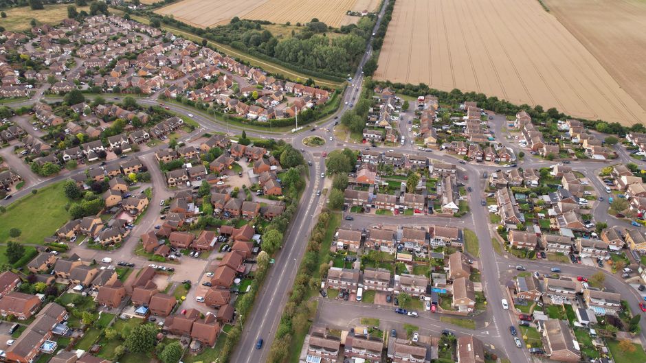 An aerial view of a residential neighbourhood showing a mix of detached, semi-detached, and terraced houses with tiled roofs, arranged along curving streets and cul-de-sacs. The image captures a central road leading through the area, with parked cars lining the streets. Adjacent to the houses are green lawns, gardens, and trees. In the background, there are large open fields with crops, separated from the residential zone by a line of trees and a main road. The lighting suggests daytime with natural light evenly illuminating the scene. The overall setting reflects a typical suburban environment suitable for home relocation and furniture transport, and the scene implies the context of moving services such as packing, loading, and transport carried out by companies like Man with Van Hither Green.