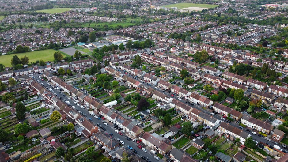 An aerial view of a residential area in Hither Green, Lewisham, showing a row of terraced houses with small front gardens, some with driveways and parked cars along the street. The image depicts the loading process for house removals, with several cardboard boxes, plastic-wrapped furniture, and packing materials on the pavement and inside the properties, ready for transportation. A van from Man with Van Hither Green is parked on the street near the entrance of one property, with its back doors open, indicating ongoing furniture transport and home relocation activities. The surrounding environment includes lush trees, green fields beyond the urban area, and a mix of other residential streets, typical of a suburban neighbourhood prepared for a house move as part of the Lewisham Council permit requirements for Hither Green removals.