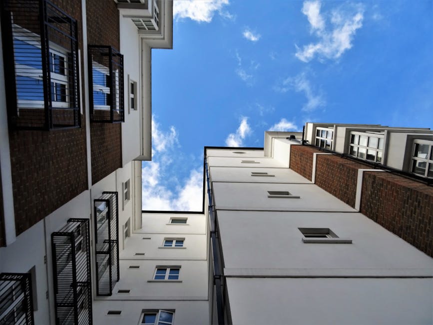 Looking upward between narrow, multi-storey residential buildings inside a limited-access flat complex on Hither Green estate, showing white facades with a few small windows and black metal balcony railings on one side. The photo captures the open sky with scattered white clouds above, indicating daylight. The scene depicts a typical urban environment where moving companies like Man with Van Hither Green often operate, assisting with home relocation and furniture transport. This perspective highlights the confined space around the buildings, which may require careful planning for packing and loading during a moving service, especially in tight courtyards or shared access areas. The arrangement of the buildings suggests the need for precise logistics when delivering packing materials, boxes, and loaded trolleys or carts to facilitate the loading process for house removals from such structures, ensuring safe transport of belongings from inside the flats to waiting vans.
