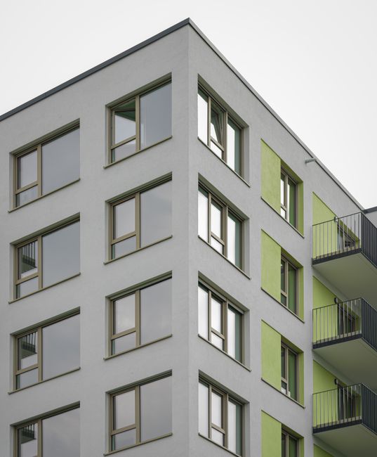 A multi-storey modern residential building with a white facade featuring multiple rectangular windows with green accents alongside grey balcony railings visible on the right side. The building is set against an overcast sky, with no visible entrance or surrounding environment. The image focuses solely on the exterior structure, highlighting its architectural design and window placement, suitable for illustrating housing or apartment relocation services related to home removals on Hither Green estate. Man with Van Hither Green may be involved in moving or packing services for such properties, supporting solutions for limited-access flats and furniture transport considerations in urban settings.
