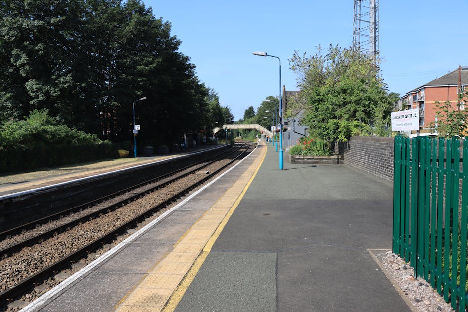 A clear view of a train station platform in daylight with dark railway tracks running parallel to it. The platform surface is made of asphalt with a yellow tactile paving strip near the edge for safety, and blue-painted lampposts line the walkway. On the right side, there is a green metal fence adjacent to a brick wall, with a small garden bed featuring some plants and flowers. Further along the platform, a white sign with black text is attached to a pole, and a covered waiting shelter with seating is visible in the background. The scene appears calm and organized, with trees and residential buildings visible beyond the platform, representing a typical suburban train station environment suitable for home relocation or furniture transport logistics managed by [COMPANY_NAME].
