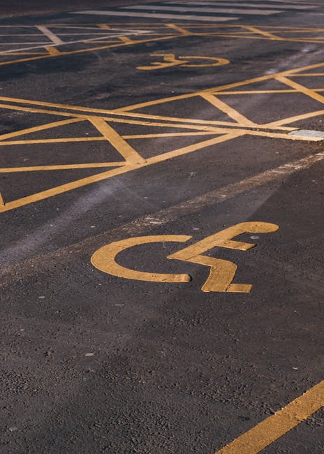 A close-up view of a paved parking area featuring clearly marked yellow wheelchair accessible parking spaces with the universal wheelchair symbol painted on the dark asphalt surface. The spaces are outlined with yellow lines, some intersecting at angles, and are situated adjacent to each other. The area appears to be outdoors with natural lighting, and the ground shows signs of wear with slight tire marks and asphalt patches. This parking lot is part of a building's exterior, potentially near a residential or commercial property, supporting accessibility during a house removal or relocation process. The image emphasizes organized parking arrangements for vehicles, relevant to the loading and unloading phases of home relocation, and is associated with [manwithvanhithergreen.co.uk] during a moving service in the SE13 area, specifically related to parking permits, loading bays, and efficient furniture transport for an internal or local move.