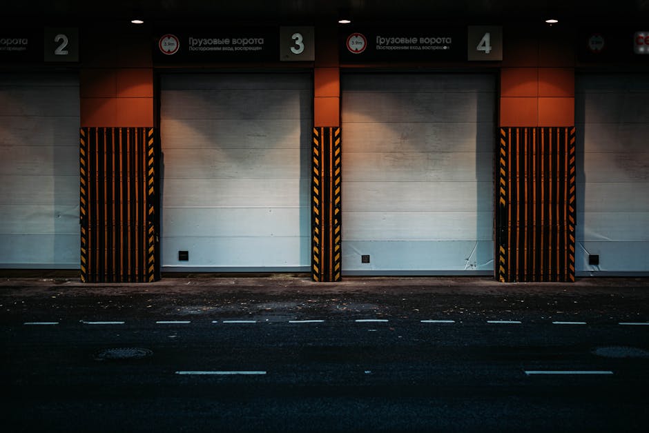 A row of three closed industrial loading bay doors in a modern building exterior, with each door numbered 2, 3, and 4. The doors are set within a white wall with beige horizontal panels. Protective black and yellow striped safety barriers are positioned in front of each door, extending from the ground to just below the ceiling to prevent vehicle impact. The area is illuminated by overhead lighting, casting subtle shadows on the surface. In front of the building, there is an asphalt loading zone with visible lane markings and some tire marks and debris. This scene depicts the typical environment where professional removals companies, such as Man with Van Hither Green, might load and unload household or office furniture during a home relocation or furniture transport process, supporting efficient parking, permits, and loading bay operations as outlined in the SE13 Moving Guide.