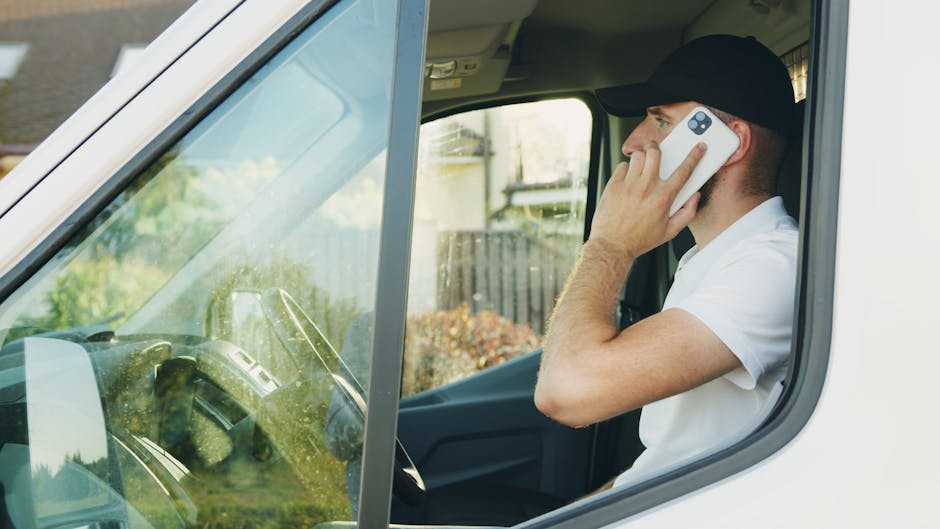 A young man with dark hair, beard, and light skin, standing outdoors in front of a white van with an open sliding door during daylight. He is wearing a light beige, long-sleeved button-up shirt with t