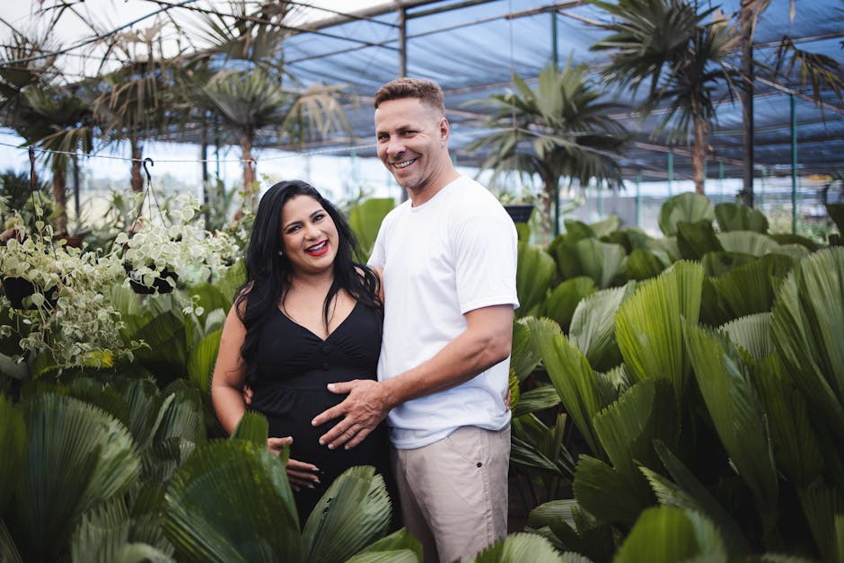 A smiling couple standing closely together inside a greenhouse filled with lush, large green palm leaves and smaller flowering plants. The woman, visibly pregnant, is wearing a black sleeveless top, while the man is dressed in a white t-shirt and beige trousers. They are positioned near the center of the image, with the man gently resting his hand on the woman's belly, both looking towards the camera. The background shows a glass or plastic roof supported by metal beams, allowing natural light to illuminate the vibrant foliage. The scene depicts a moment of connection and happiness amid the greenery, with the environment suggesting a plant nursery or botanical setting. This image is associated with house removals and moving services, highlighting themes of packing, transport, or home relocation between plants and furniture.