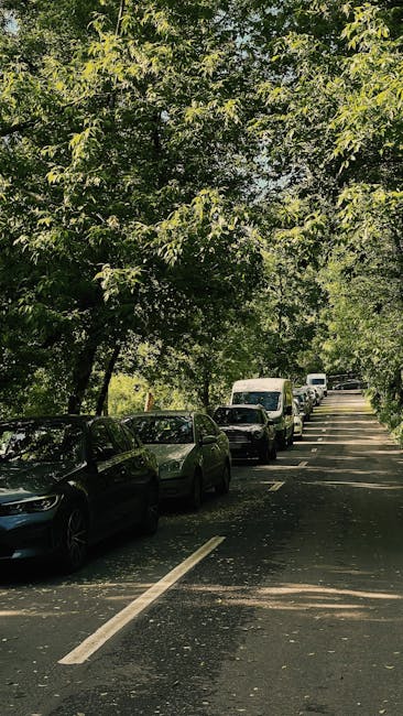 A street scene featuring a narrow residential road lined with parked cars on both sides, with a canopy of green trees extending over the street, casting dappled sunlight onto the pavement. Several vehicles, including a white van, are visible further down the street, which is typical for a quiet urban or suburban neighbourhood. The road appears to be part of a house relocation or moving process, as suggested by the context of [COMPANY_NAME], with the setting showing the environment where furniture transport and packing services might be coordinated during a home move in Hither Green. No moving equipment or furniture is visible in this image, but the overall environment provides the context of a street suitable for a professional removals company's operations, such as loading or unloading in preparation for a vehicle-based home relocation.